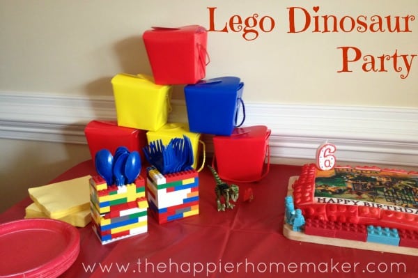 A collection of dinosaur lego party including bins, silverware holders made of legos and a birthday cake