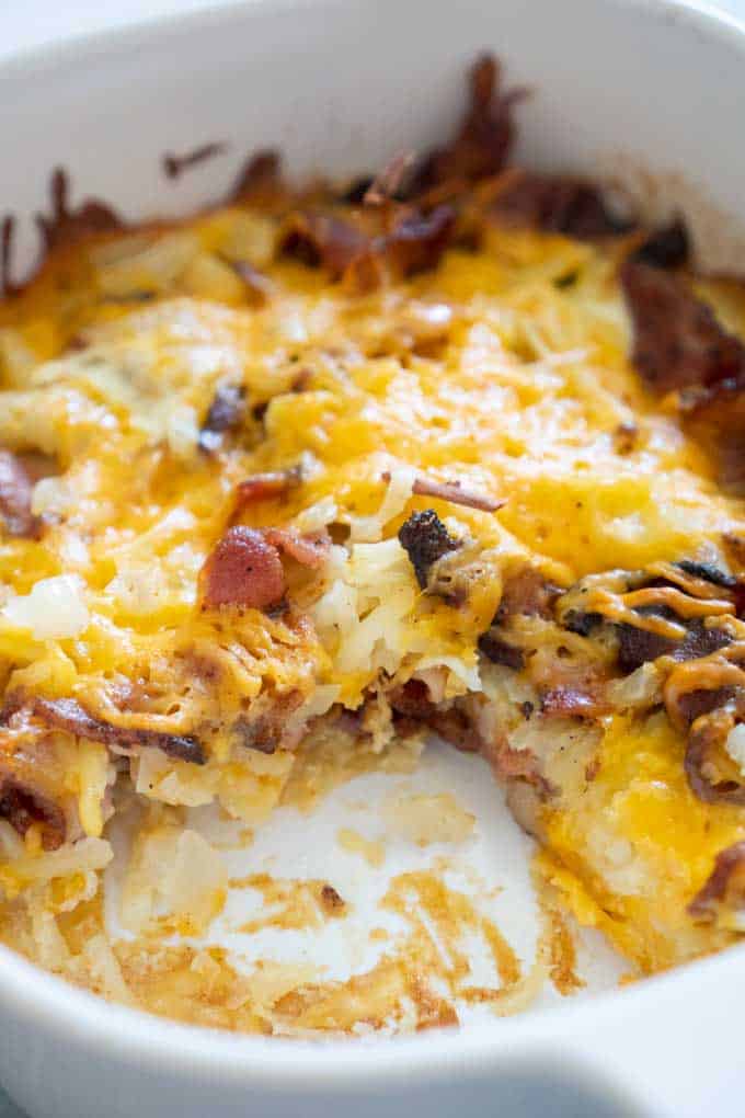 cheesy bacon hash brown casserole in white casserole dish with scoop missing