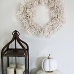 A DIY giant pompom wreath hanging over a white pumpkin and lantern