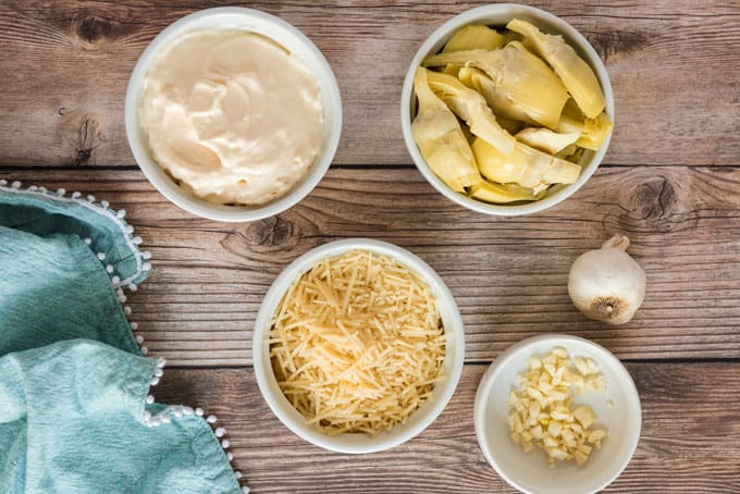 overhead view of white bowls with mayonnaise, parmesan cheese, artichoke hearts and garlic for dip recipe