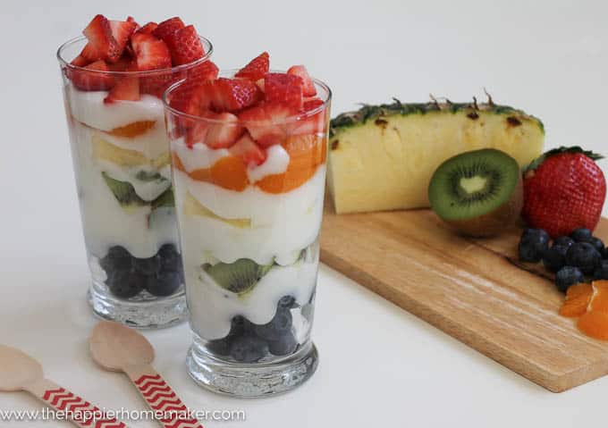 A close up of two glasses of rainbow yogurt parfaits topped with strawberries with a cut pineapple and kiwi on a cutting board in the background