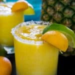 A close up of a frozen pineapple margarita garnished with a slice of lime and lemon in front of a pineapple
