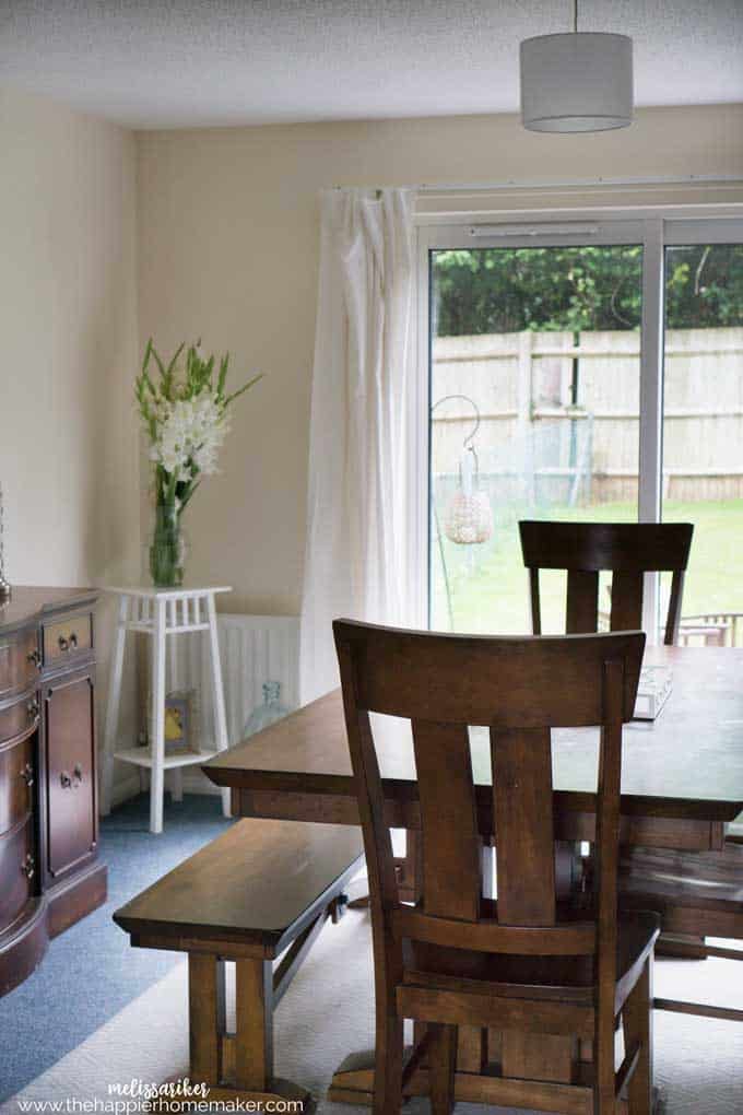 A brown dining room table with a white plant stand and flowers in the background