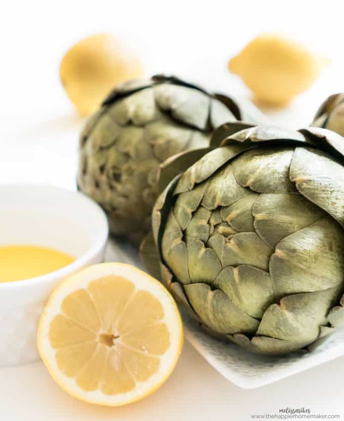 A close up of two artichokes with a halved lemon and butter sauce