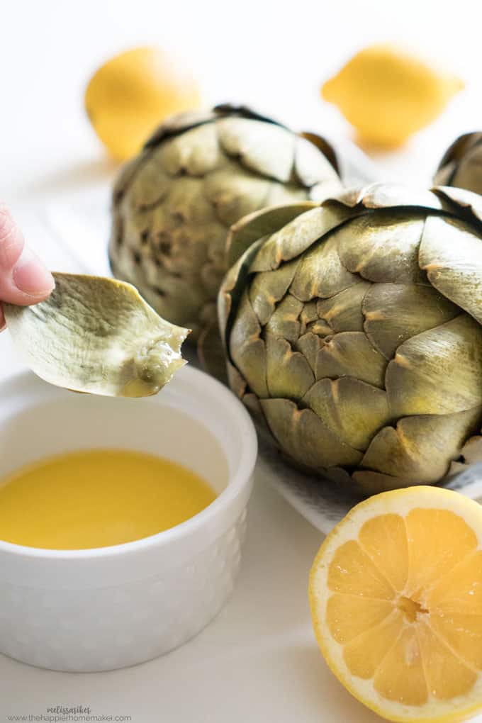 A close up of two artichokes on a white plate with butter sauce, halved lemon showing a leaf dipped in butter