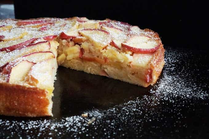 A close up of a French apple cake after baking with a slice removed to show the inside of the cake on a dark table with powdered sugar around it