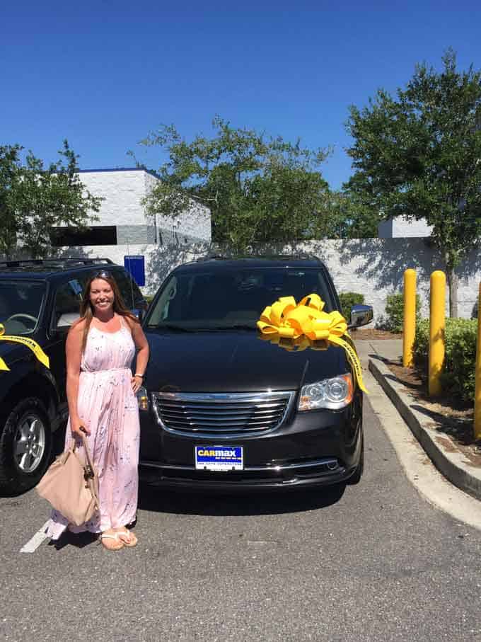 A woman in a dress standing in front of a new minivan with a yellow bow on the minivan 