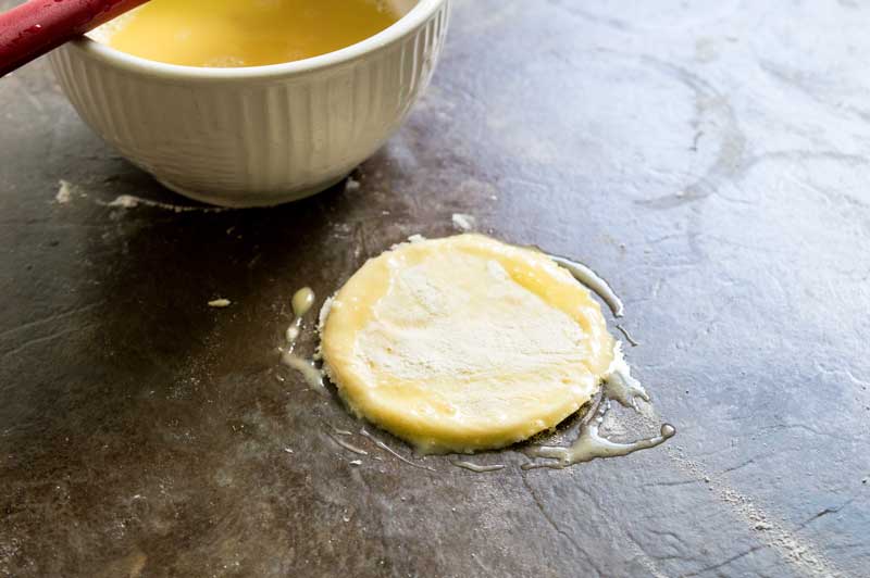 egg wash on three inch circle of puff pastry dough on black baking sheet with ramekin of egg wash for apple hand pies