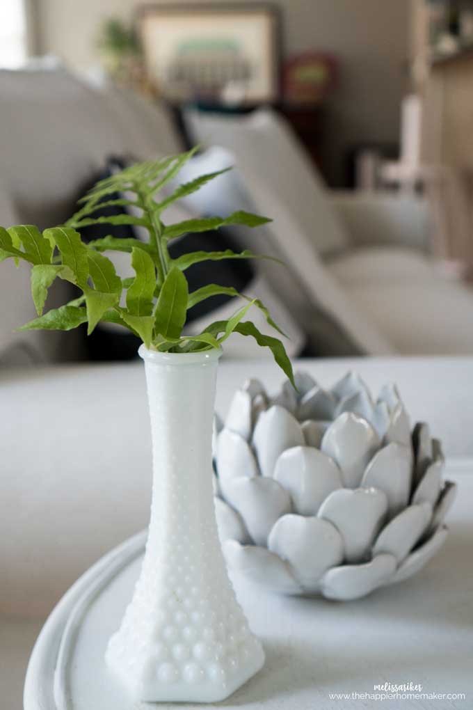A close up of a fern leaf sitting in a white vase with a ceramic white avocado behind it