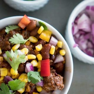 A bowl of chili garnished with fresh corn, cilantro, onion and red pepper in a white bowl