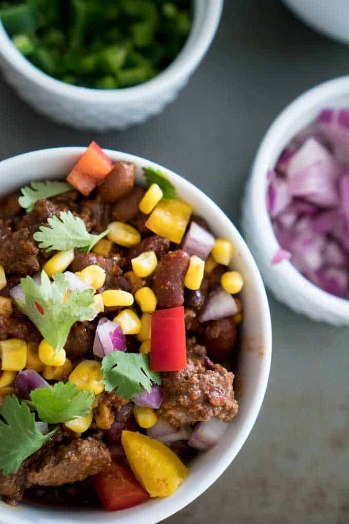 A bowl of chili garnished with fresh corn, cilantro, onion and red pepper in a white bowl