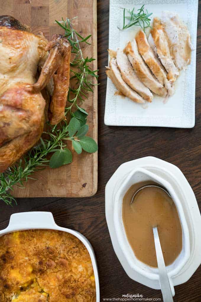 Een gekookte kalkoen op een snijplank gegarneerd met groen naast gesneden kalkoen op een witte schotel met jus en een braadpan voor Thanksgiving