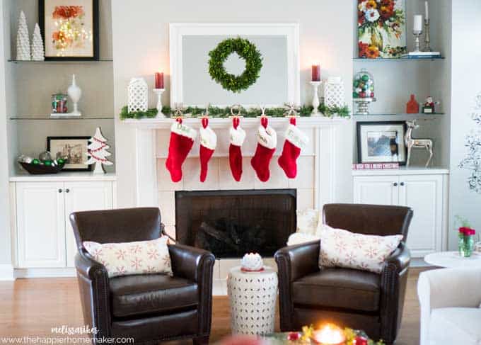 A living room decorated for Christmas with stockings hanging from the mantel, a mirror with wreath, and two leather chairs in the foreground 