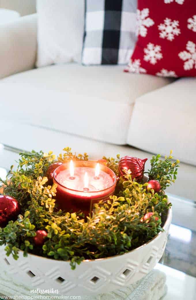 A close up of a red holiday candle surrounded by Christmas ornaments and faux box wood garland