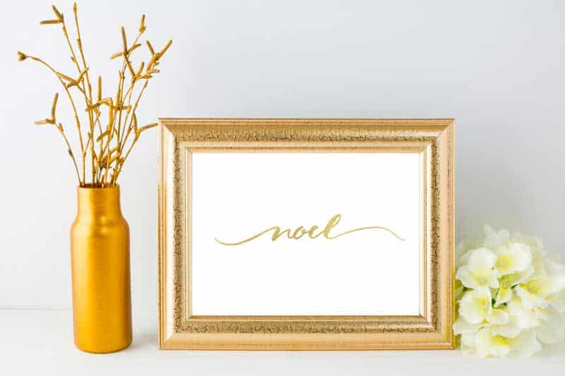 A gold picture frame with the word Noel written in calligraphy next to a gold vase of wheat