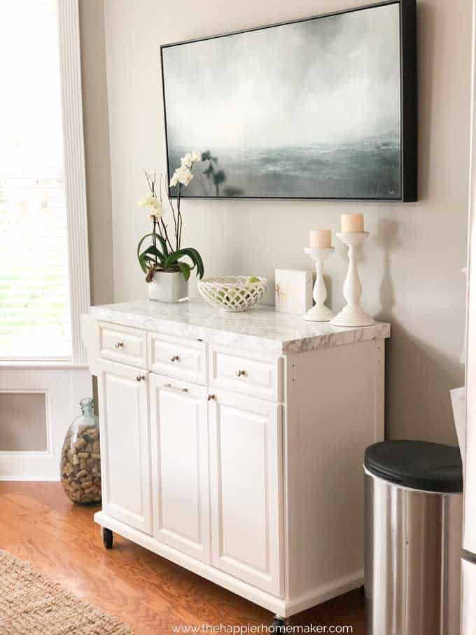 white rolling kitchen island with marble top below a large ocean painting