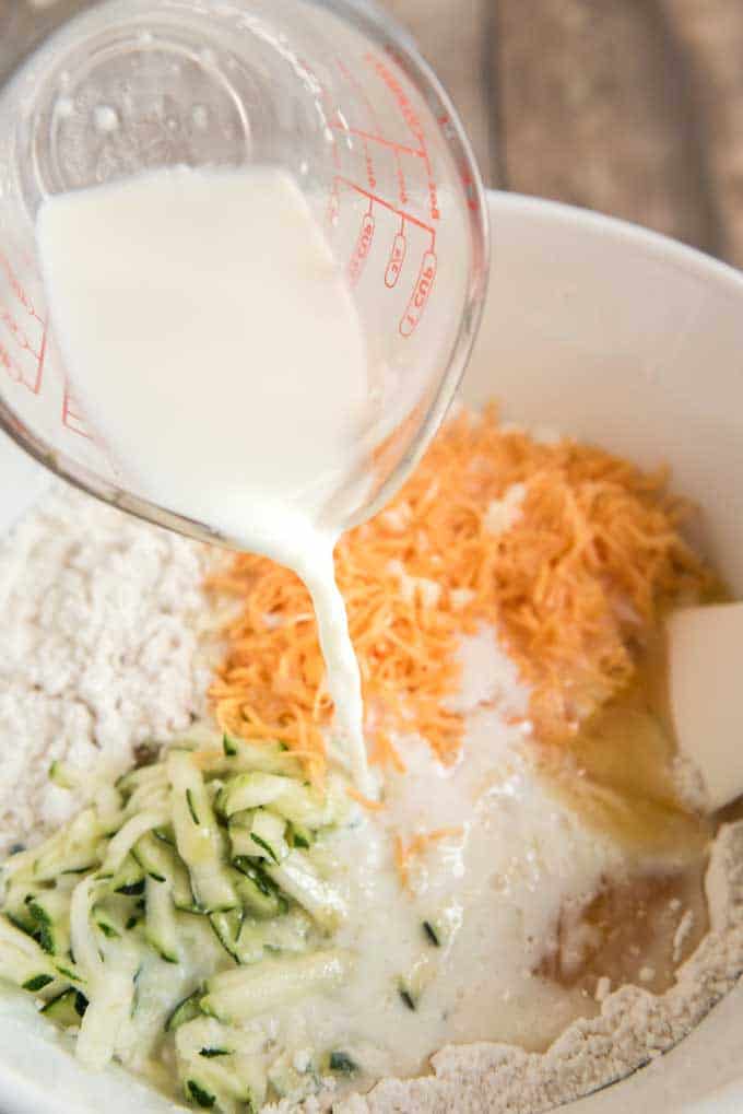 pouring milk out of glass measuring cup into white bowl with cheese, shredded zucchini and flour for cheddar zucchini bread