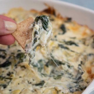 Hand holding cracker scooping up Spinach Artichoke Dip from white casserole dish