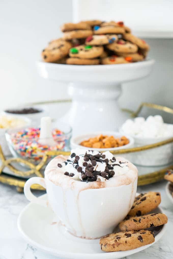 hot chocolate in white mug with whipped cream and sprinkles and cookies on cake stand in the background