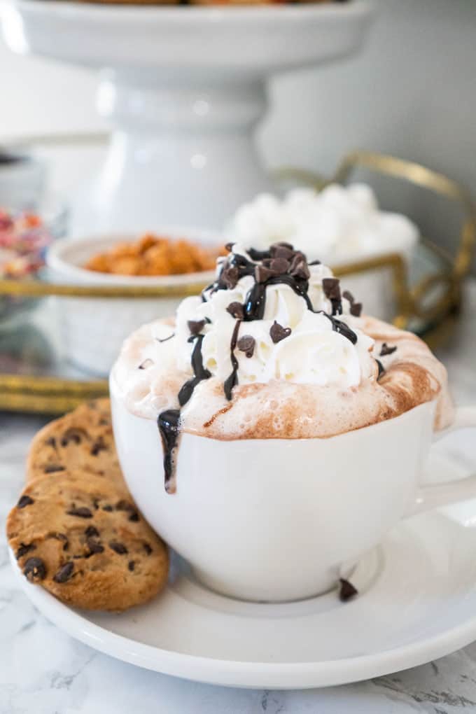 hot chocolate in white mug with whipped cream and chocolate syrup and chocolate chips and cookies on the saucer below