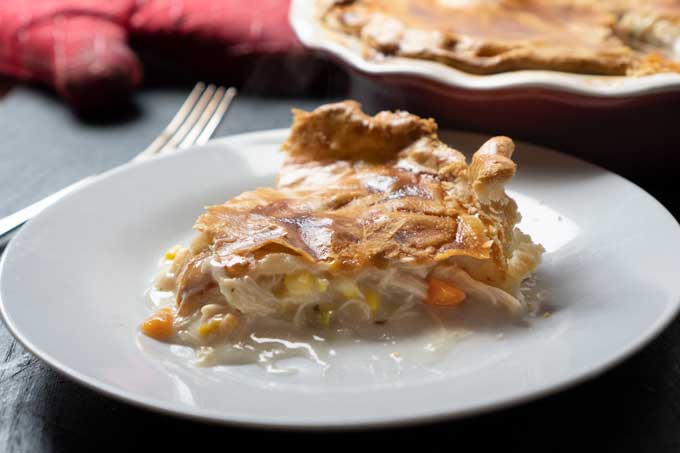 slice of homemade chicken pot pie on white plate with whole pie in background along with fork