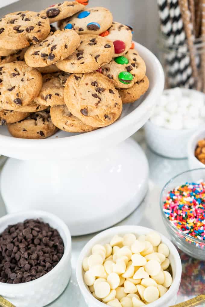 hot chocolate bar with cookies stacked on cake stand and various toppings in small bowls surrounding