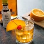 old fashioned bourbon cocktail with orange slice and cherry in front of bottle of bitters and sliced orange on cutting board