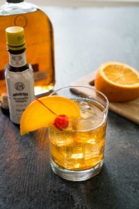 old fashioned bourbon cocktail with orange slice and cherry in front of bottle of bitters and sliced orange on cutting board