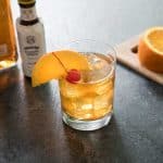 old fashioned bourbon cocktail with bottle of bitters and garnished with orange slice and cherry