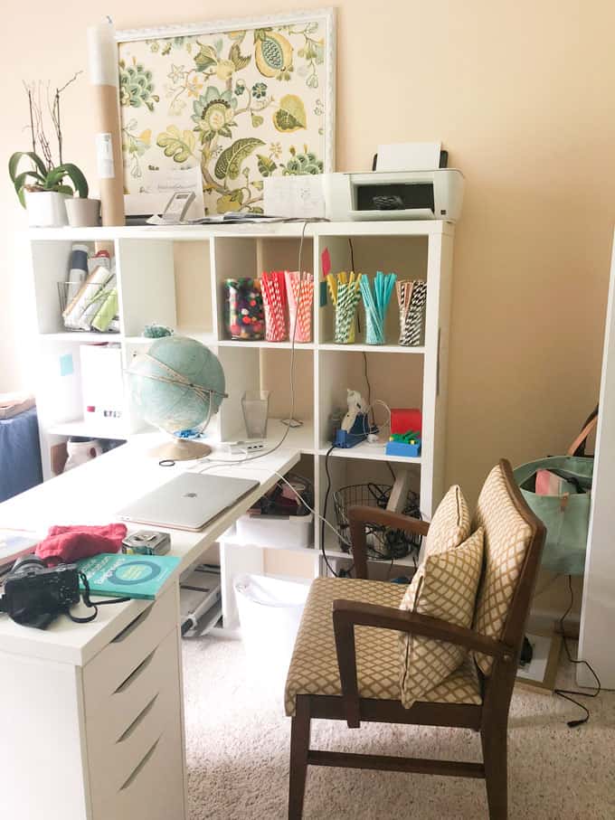 cluttered office with white cube style book shelves and white ikea desk and midcentury vintage chair