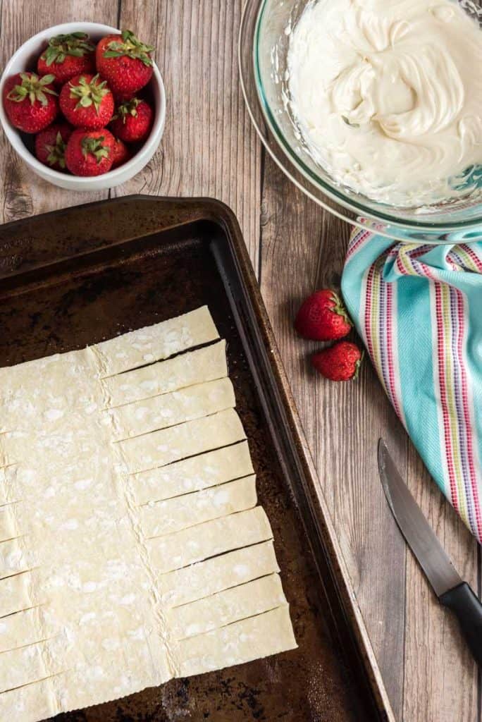 puff pastry dough on cookie sheet with strips cut out to braid a danish with bowl of strawberries and colorful napkin nearby