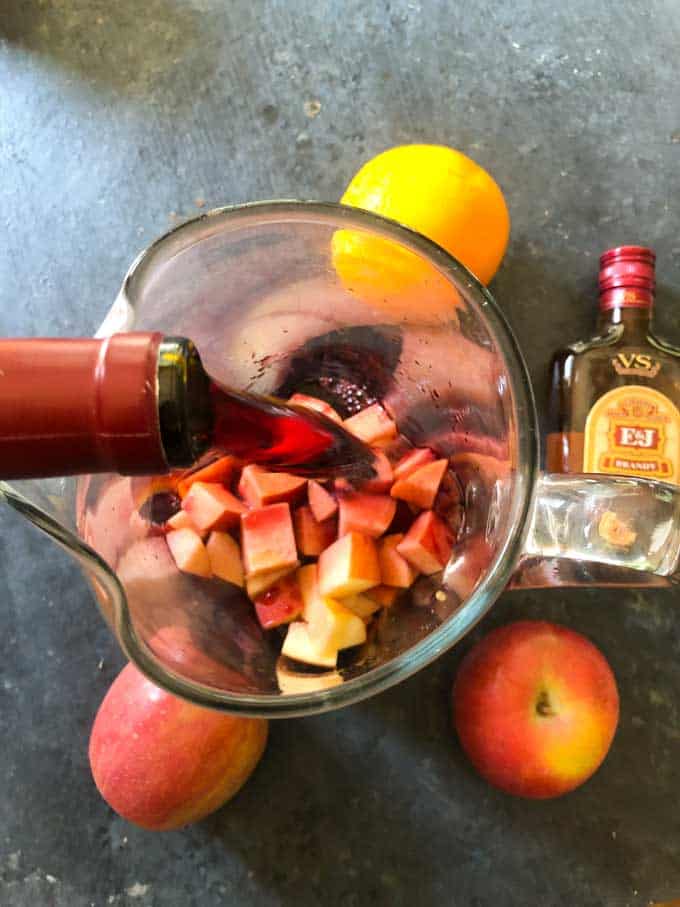 pouring red wine into glass pitcher with chopped fruit to make sangria