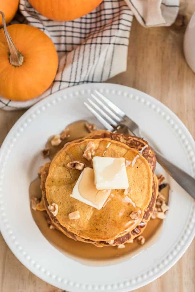 overhead view of pumpkin pancakes with butter and syrup on white plate with fork and small pumpkin on wood table