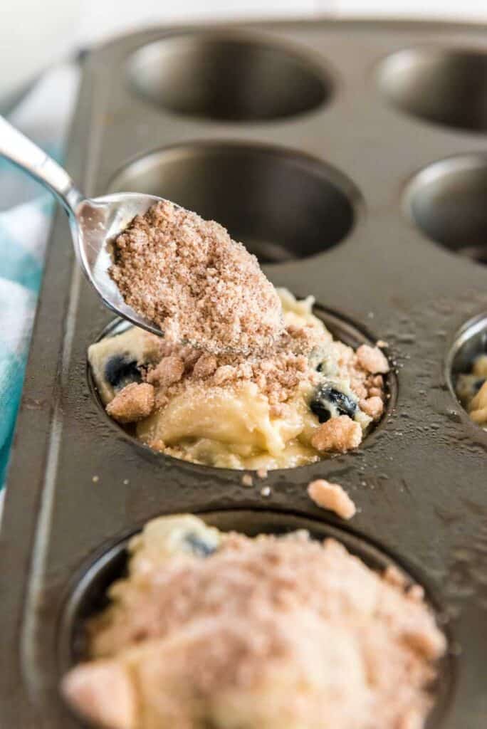 spoon adding crumble topping to blueberry muffin batter in pan