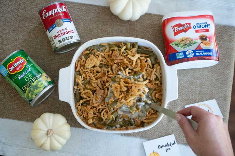 green bean casserole in white dish surrounded by canned cream of mushroom soup, green beans, crispy onions and white small pumpkins