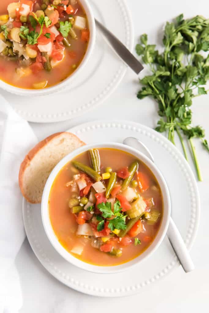 vegetable soup in two white bowls on a white plates with slice of bread and butter knife on white table with fresh parsley bunch