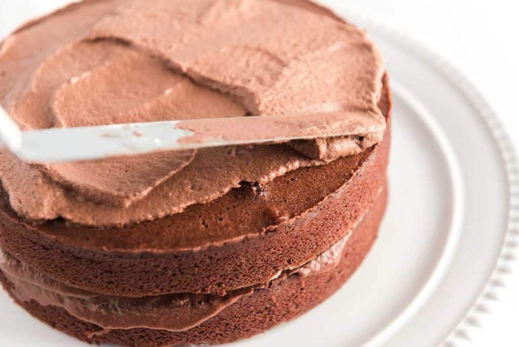 chocolate cake being iced with chocolate frosting on white cake stand