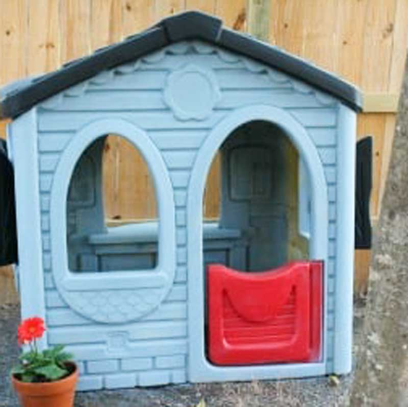 plastic playhouse painted blue wit black roof and red door in front of wood fence