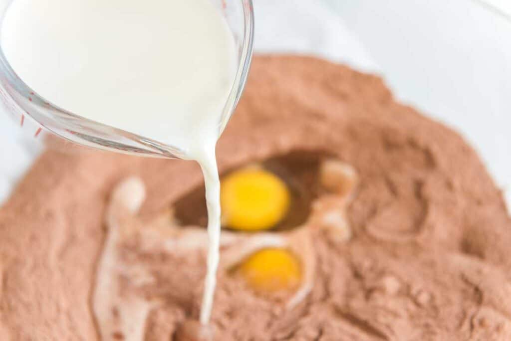 pouring milk into bowl with cocoa powder and eggs for chocolate cake