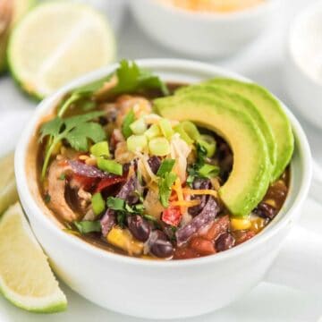 slow cooker chicken tortilla soup in white bowl with avocado slices and cilantro on top and lime in background