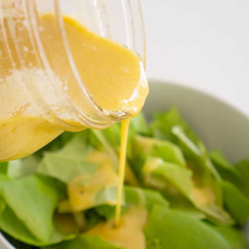 dijon salad dressing pouring onto greens in white bowl