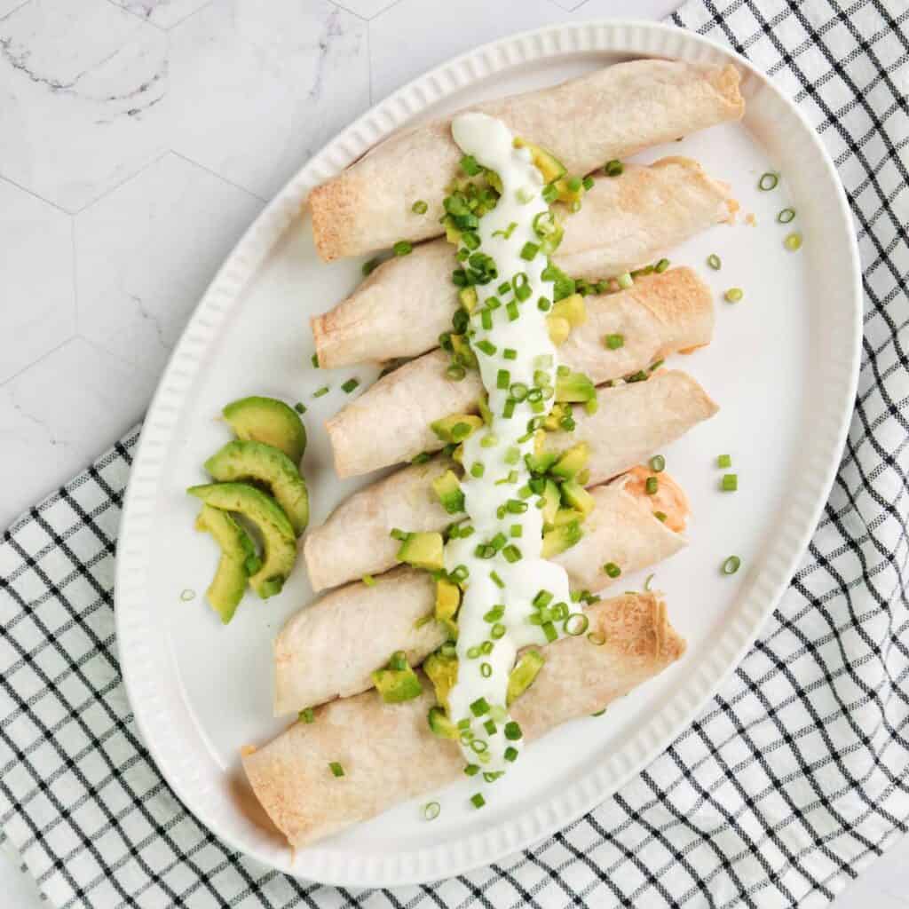 overhead view of platter with chicken and cheese taquitos topped with sour cream, avocado, and green onions
