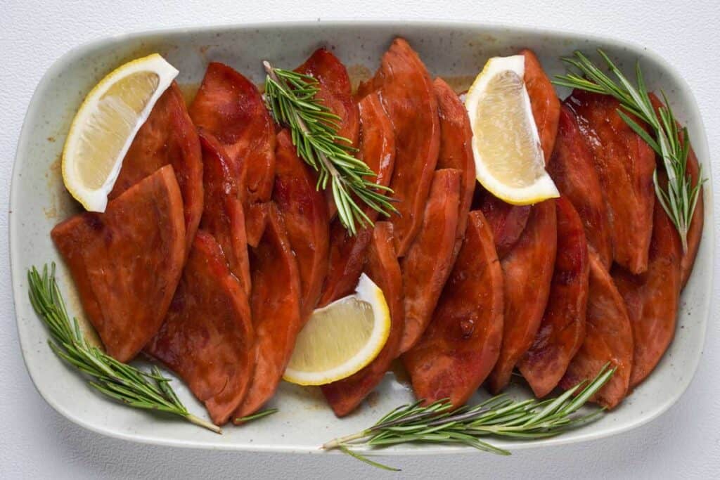 plate with sliced glazed ham garnished with rosemary and lemon wedges
