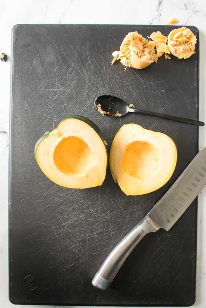halved acorn squash with seeds scooped out on cutting board with knife and spoon