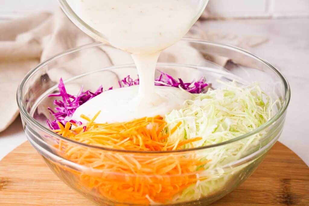 pouring coleslaw dressing over shredded cabbage and carrots